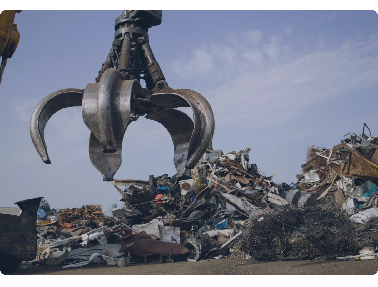 An orange-peel grapple hanging over a mound of mixed scrap metal under a blue sky.