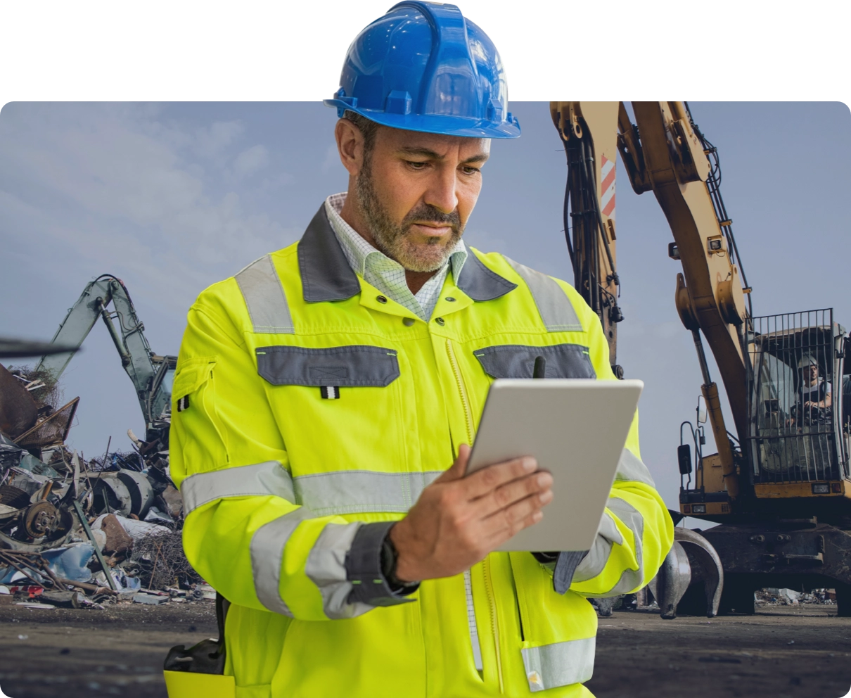 A yard worker in a blue hard hat and hi-vis jacket reading a tablet, with excavators and scrap piles behind him.