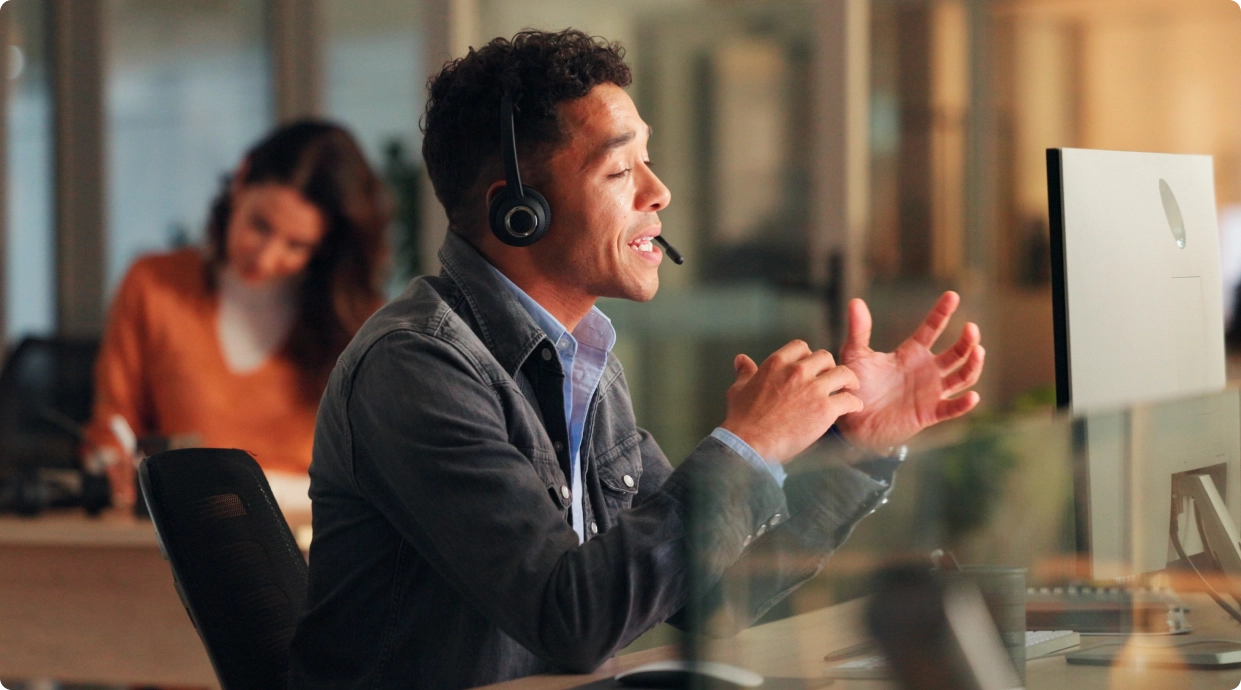 A support agent wearing a headset talking at his desk in a softly lit office, a colleague working behind him.