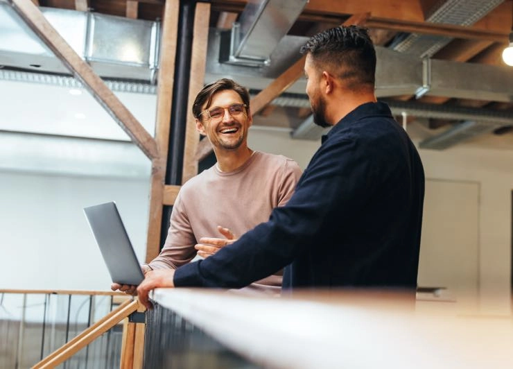 Two colleagues talking at a railing in a loft office, one holding a laptop.