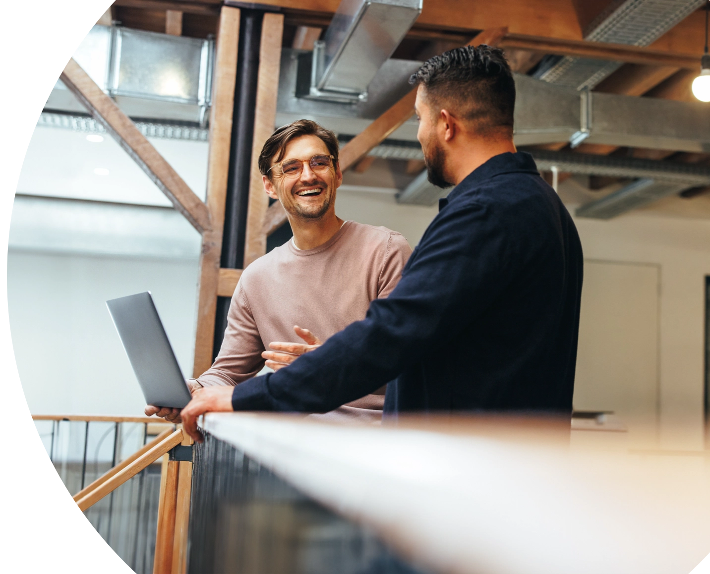 Two colleagues talking at a railing in a loft office, one holding a laptop.