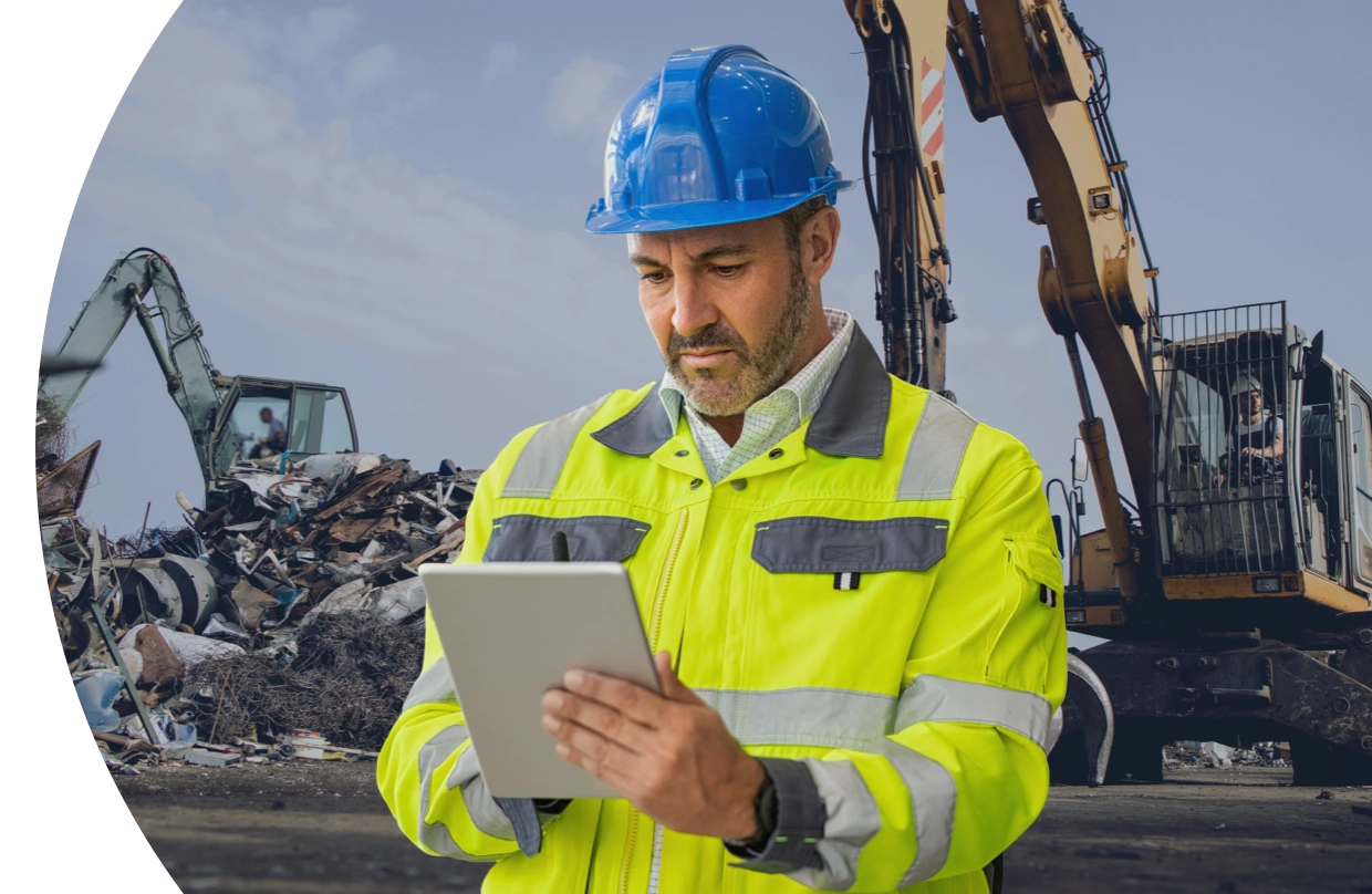 A yard worker in a hard hat and hi-vis jacket entering a ticket on a tablet beside a loaded scrap pile.